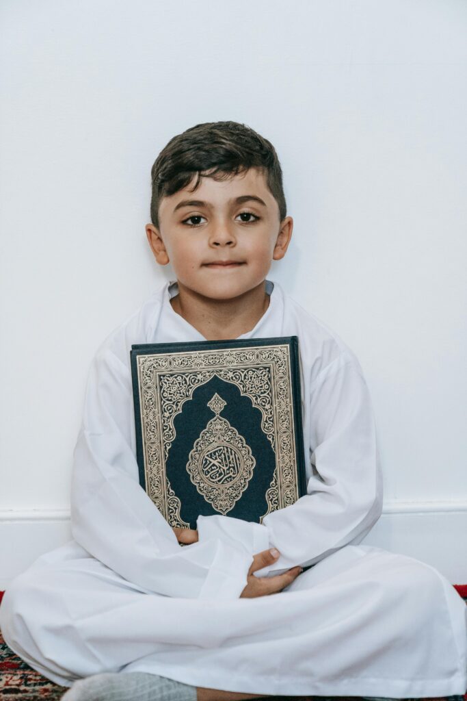 A young boy sitting indoors, holding the Koran in traditional white attire.